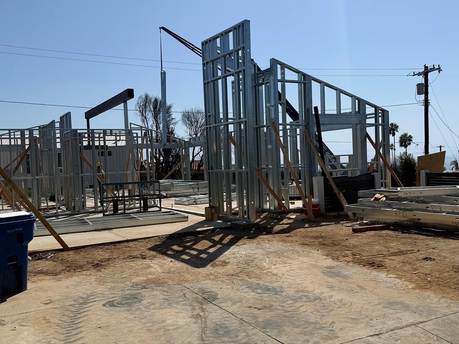 The site early on: the first walls stand, the crane sets the next, and numbered panels wait on the right. Job site in Pacific Palisades with galvanized steel wall panels standing on a slab, a crane boom behind them and numbered panels stacked on the right
